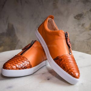Könnte beinhalten: Orangefarbene Ledersneaker mit Reißverschluss und weißen Sohlen. Zehen und Ferse weisen ein Schuppenmuster auf. Die Schuhe werden auf einer Marmoroberfläche präsentiert.