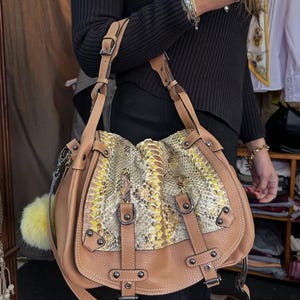 Könnte beinhalten: Hellbraune Lederhandtasche mit Schlangenmuster auf der oberen Klappe. Die Tasche hat einen langen Schulterriemen, mehrere Schnallen und einen gelben Pompon-Anhänger. Die Tasche ist im Satchel-Stil.