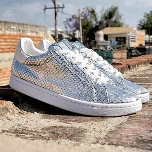 Könnte beinhalten: Ein Paar silberne Sneaker mit einem strukturierten, schuppenartigen Muster. Die Schuhe haben weiße Schnürsenkel, eine weiße Zunge und eine weiße Sohle. Die Sneaker werden im Freien auf einer Ziegeloberfläche präsentiert.
