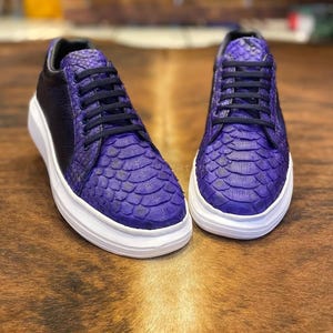 Könnte beinhalten: Ein Paar lila Sneaker mit einem strukturierten, schuppenartigen Muster auf Zehen und Seiten. Die Schuhe haben schwarze Schnürsenkel und eine schwarze Fersenpartie, die auf einer weißen Sohle sitzen. Die Sneaker stehen auf einer braunen Oberfläche.