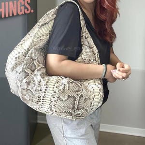 Può includere: Una grande borsa tote beige e marrone con motivo a pelle di serpente e un'ampia tracolla. La borsa è tenuta da una persona che indossa una maglietta nera e pantaloni della tuta grigi. Il testo "WaoneLeather" è visibile.