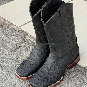 May include: A pair of black cowboy boots with a square toe and a textured, snake-like pattern on the lower part. The upper part of the boots features decorative stitching. The boots have a brown sole and a white trim.