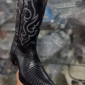May include: Black cowboy boot with a textured lizard skin pattern. The boot features decorative stitching in a lighter color along the shaft. The toe is pointed, and the heel is angled. The boot is held by a person.