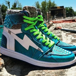 Pu&ograve; includere: Sneakers alte color teal con motivo a pelle di serpente, dettagli bianchi e lacci verde neon. Le scarpe presentano un fulmine bianco sul lato e una suola bianca. Le sneakers sono esposte all'aperto.