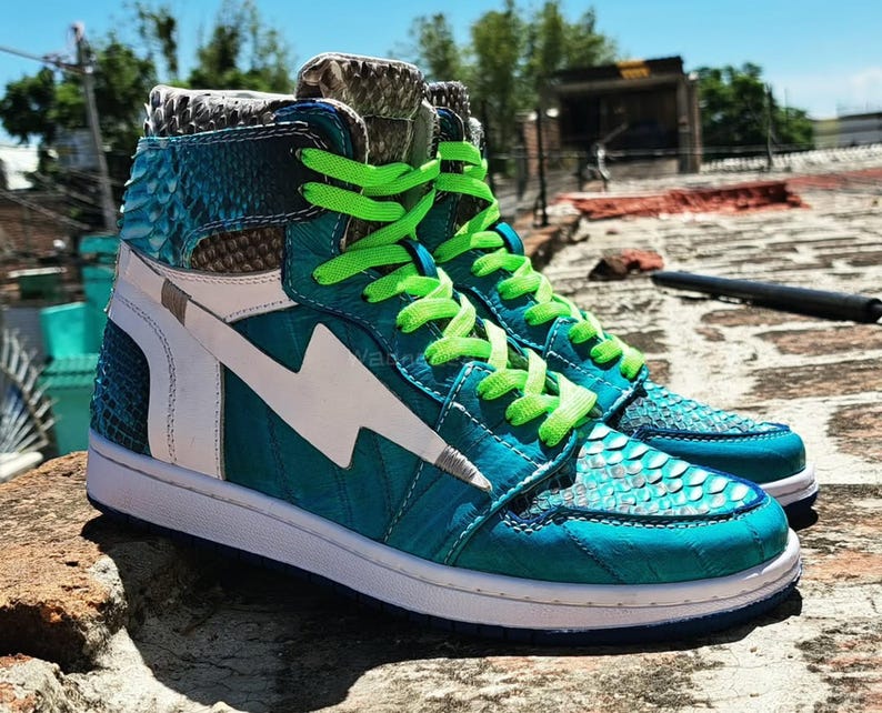 Pu&ograve; includere: Sneakers alte color teal con motivo a pelle di serpente, dettagli bianchi e lacci verde neon. Le scarpe presentano un fulmine bianco sul lato e una suola bianca. Le sneakers sono esposte all'aperto.