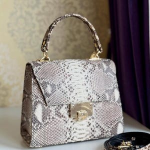 Op de afbeelding: Handtas met een slangenprint in grijstinten, wit en bruin. De tas heeft een handvat aan de bovenkant en een goudkleurige sluiting. Er is ook een bijpassende slangenleren riem aanwezig.