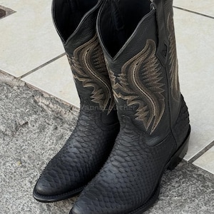 Könnte beinhalten: Ein Paar schwarze Cowboy-Stiefel mit aufwendiger goldfarbener Stickerei am Schaft. Die Stiefel haben ein strukturiertes, schlangenartiges Muster am unteren Teil und eine spitze Zehenpartie. Die Stiefel sind aus Leder gefertigt.
