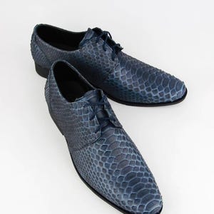 May include: A pair of blue leather dress shoes with a textured, snakeskin-like pattern. The shoes feature a classic lace-up design and a low heel. The colour is a deep, mottled blue, and they are set against a white background.