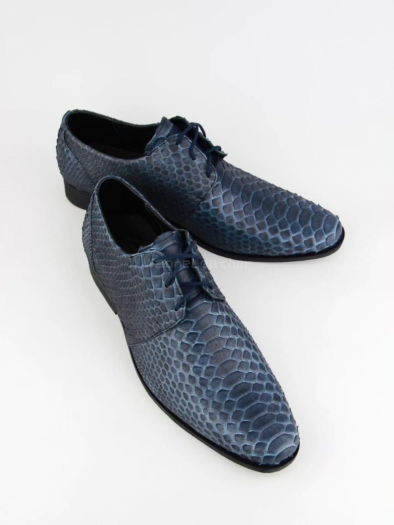May include: A pair of blue leather dress shoes with a textured, snakeskin-like pattern. The shoes feature a classic lace-up design and a low heel. The colour is a deep, mottled blue, and they are set against a white background.