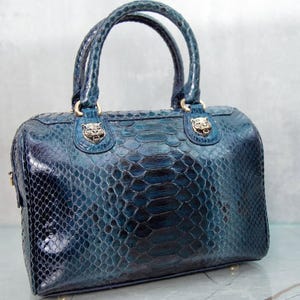 Pode incluir: Bolsa de mão azul-petróleo com padrão de pele de cobra. A bolsa tem duas alças trançadas e detalhes dourados. A parte superior da bolsa apresenta dois emblemas decorativos com cabeça de leão.