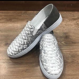 Könnte beinhalten: Ein Paar Slip-On-Sneaker mit Schlangenmuster in Grau- und Weißtönen. Die Schuhe haben weiße Sohlen und eine dunkelgraue Ferse. Die Sneaker werden auf einer Holzoberfläche präsentiert.