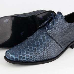 May include: Blue leather dress shoes with a textured, snakeskin-like pattern. The shoes have a lace-up design and a dark sole. The second shoe is visible in the background, with the sole facing up. The shoes are on a white background.