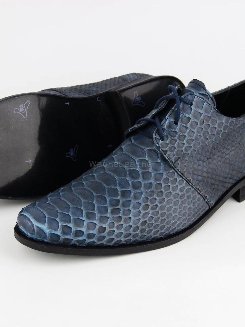 May include: Blue leather dress shoes with a textured, snakeskin-like pattern. The shoes have a lace-up design and a dark sole. The second shoe is visible in the background, with the sole facing up. The shoes are on a white background.