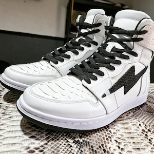 May include: Pair of white high-top sneakers with black laces. The sneakers feature a textured, scale-like pattern on the toe and side panels. A black lightning bolt design is on the side. The soles are white with a black outsole.
