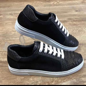 Puede incluir: Zapatillas negras con cordones y suelas blancas. La puntera y el talón del zapato tienen un patrón texturizado similar a la piel de serpiente. El cuerpo principal del zapato es de ante negro. Las zapatillas se exhiben sobre una superficie de madera.
