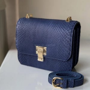 May include: A navy blue handbag with a snakeskin texture, featuring gold-coloured hardware on the clasp and strap attachments. The matching strap is coiled beside the bag. The text "WanneLeather" is visible on the bag.