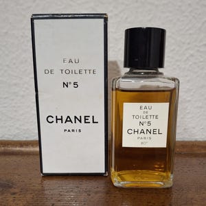 Pode incluir: Um frasco de Eau de Toilette Chanel N&deg;5 e sua caixa. A caixa e o frasco apresentam o logotipo da Chanel e as palavras "Eau de Toilette N&deg;5 PARIS". O frasco tem uma tampa preta e cont&eacute;m um l&iacute;quido &acirc;mbar claro.