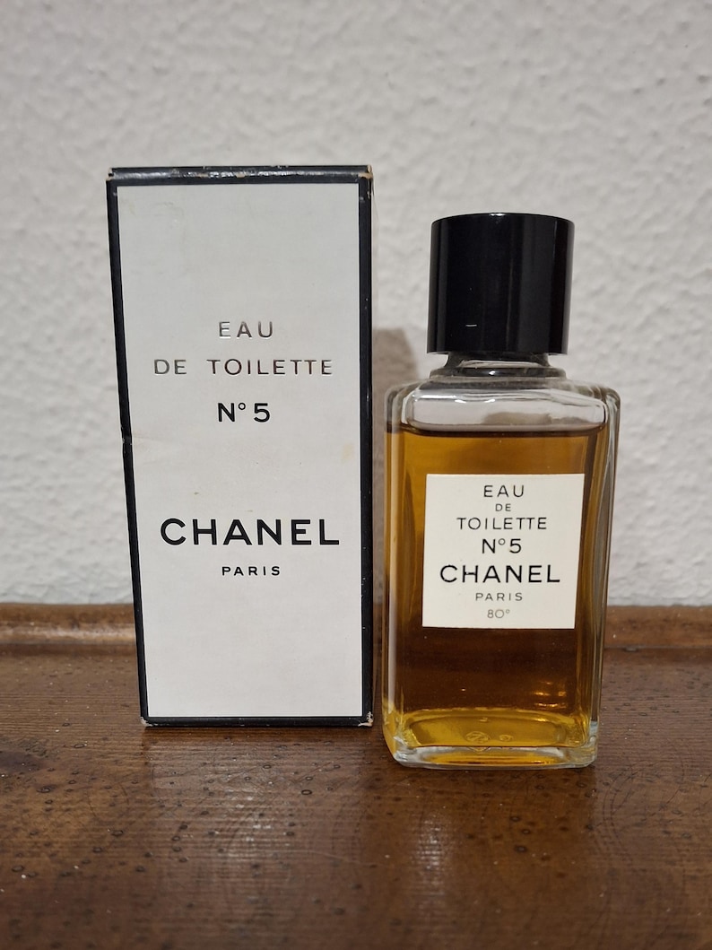Pode incluir: Um frasco de Eau de Toilette Chanel N&deg;5 e sua caixa. A caixa e o frasco apresentam o logotipo da Chanel e as palavras "Eau de Toilette N&deg;5 PARIS". O frasco tem uma tampa preta e cont&eacute;m um l&iacute;quido &acirc;mbar claro.