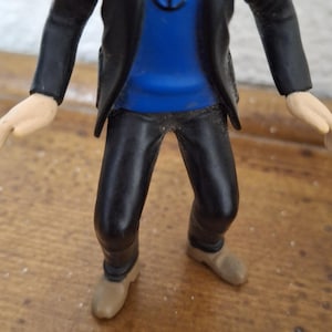 K&ouml;nnte beinhalten: Eine Kunststofffigur eines b&auml;rtigen Charakters, der eine schwarze Jacke &uuml;ber einem leuchtend blauen Pullover mit einem schwarzen Ankersymbol auf der Brust tr&auml;gt. Die Figur hat eine schwarze Hose und beige Schuhe sowie hellh&auml;utige H&auml;nde.