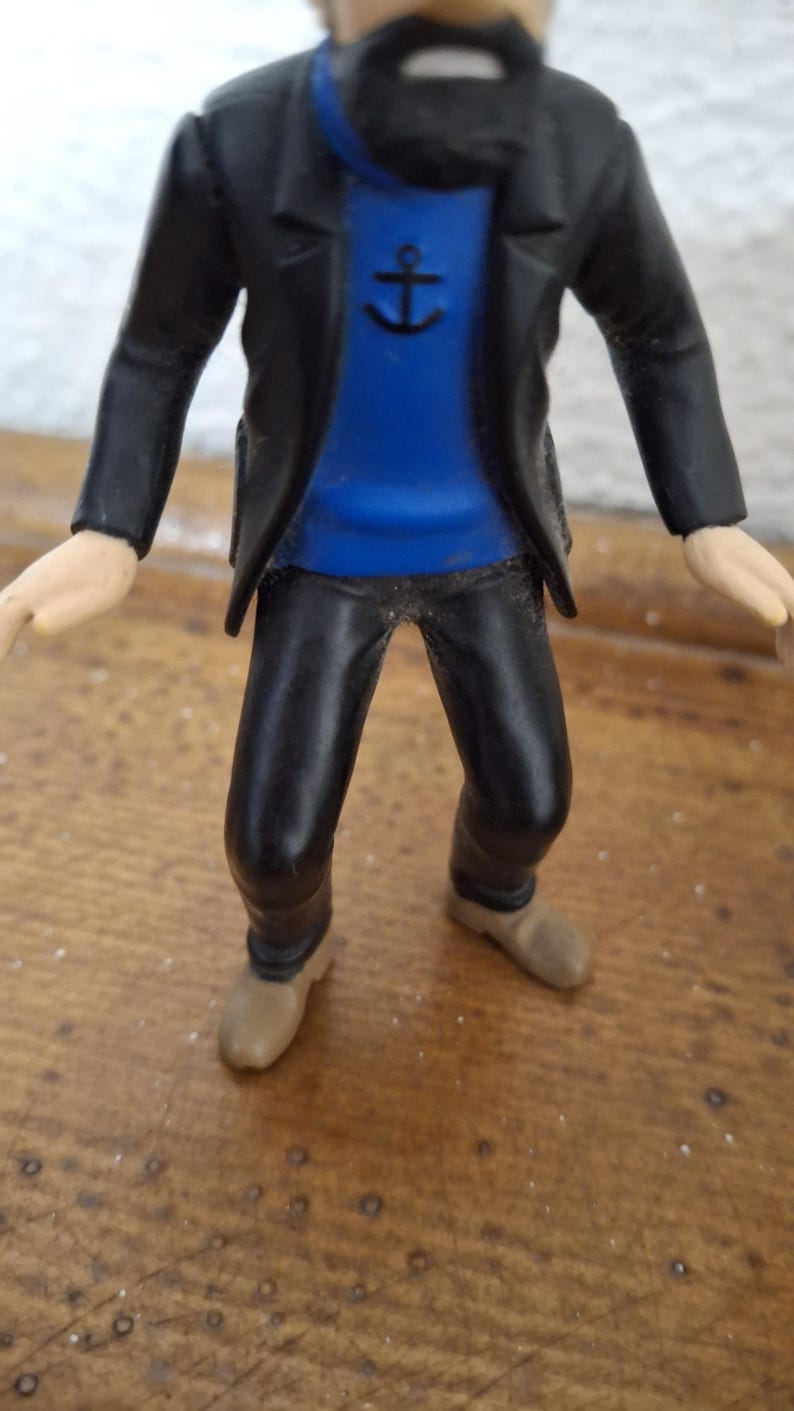 K&ouml;nnte beinhalten: Eine Kunststofffigur eines b&auml;rtigen Charakters, der eine schwarze Jacke &uuml;ber einem leuchtend blauen Pullover mit einem schwarzen Ankersymbol auf der Brust tr&auml;gt. Die Figur hat eine schwarze Hose und beige Schuhe sowie hellh&auml;utige H&auml;nde.