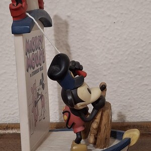 May include: A Mickey Mouse figurine in a boat, steering a wooden wheel. The figurine is part of a bookend with the text "Mickey Mouse in Steamboat Willie". The bookend has red and white accents.