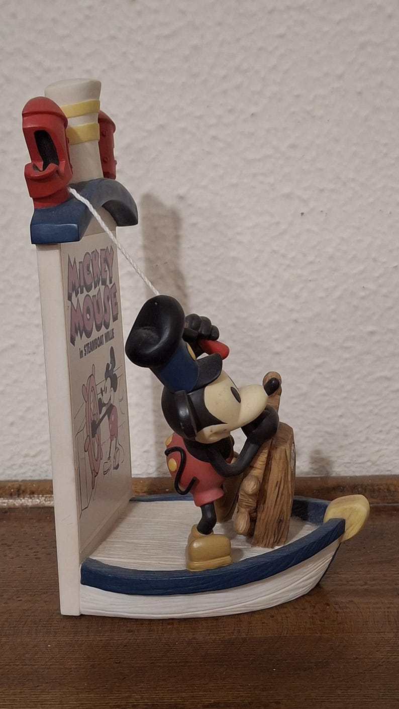 May include: A Mickey Mouse figurine in a boat, steering a wooden wheel. The figurine is part of a bookend with the text "Mickey Mouse in Steamboat Willie". The bookend has red and white accents.