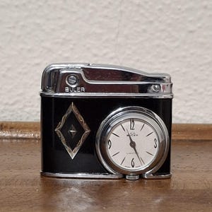 Könnte beinhalten: Ein schwarzes und silbernes EULER-Feuerzeug im Vintage-Stil mit eingebauter Uhr. Das Zifferblatt ist weiß mit schwarzen Ziffern und Zeigern. Das Feuerzeug hat ein dekoratives rautenförmiges Design. Das Wort "EULER" ist oben aufgedruckt.