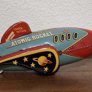 May include: Vintage tin toy rocket in teal, red, yellow, and black. The rocket has the words "ATOMIC ROCKET" and "0000" printed on the side. A lever is labeled "PUSH". The rocket has a retro space theme.