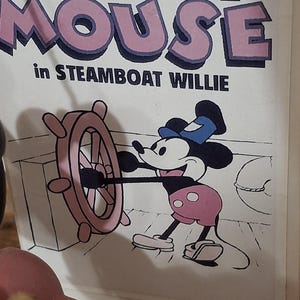 May include: A cartoon image of Mickey Mouse in Steamboat Willie. Mickey is wearing pink shorts and a blue hat, and is holding the ship's wheel. The words "Mickey Mouse" are in pink letters with a purple outline.