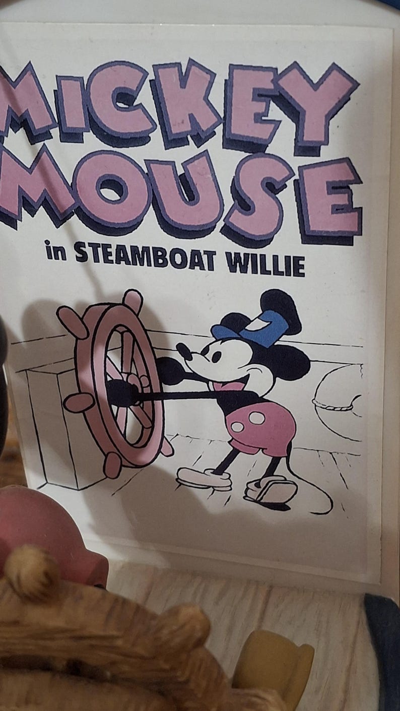 May include: A cartoon image of Mickey Mouse in Steamboat Willie. Mickey is wearing pink shorts and a blue hat, and is holding the ship's wheel. The words "Mickey Mouse" are in pink letters with a purple outline.