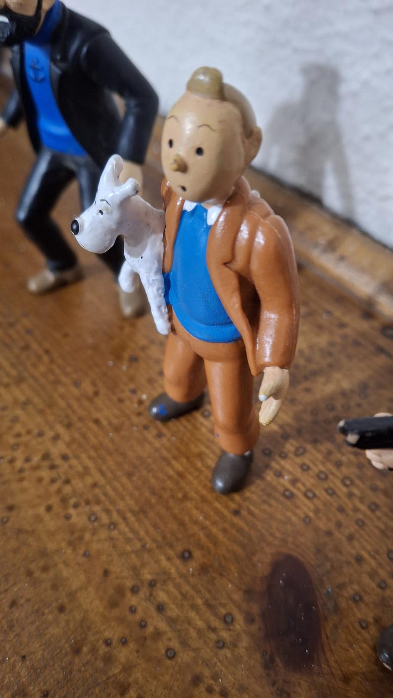 K&ouml;nnte beinhalten: Eine Kunststofffigur eines jungen Charakters mit charakteristischer Tolle, bekleidet mit einer braunen Jacke, einem blauen Pullover und einer braunen Hose, die einen kleinen wei&szlig;en Hund mit schwarzen Ohren und Nase h&auml;lt, neben einer weiteren Figur.