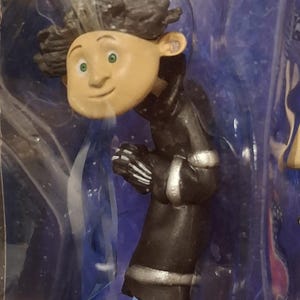 K&ouml;nnte beinhalten: Eine Figur einer Figur aus dem Animationsfilm "Coraline". Die Figur hat einen hellen Teint, gr&uuml;ne Augen und dunkles, lockiges Haar. Sie tr&auml;gt einen schwarzen Mantel mit silbernen Verzierungen, eine blaue Hose und schwarze Schuhe.