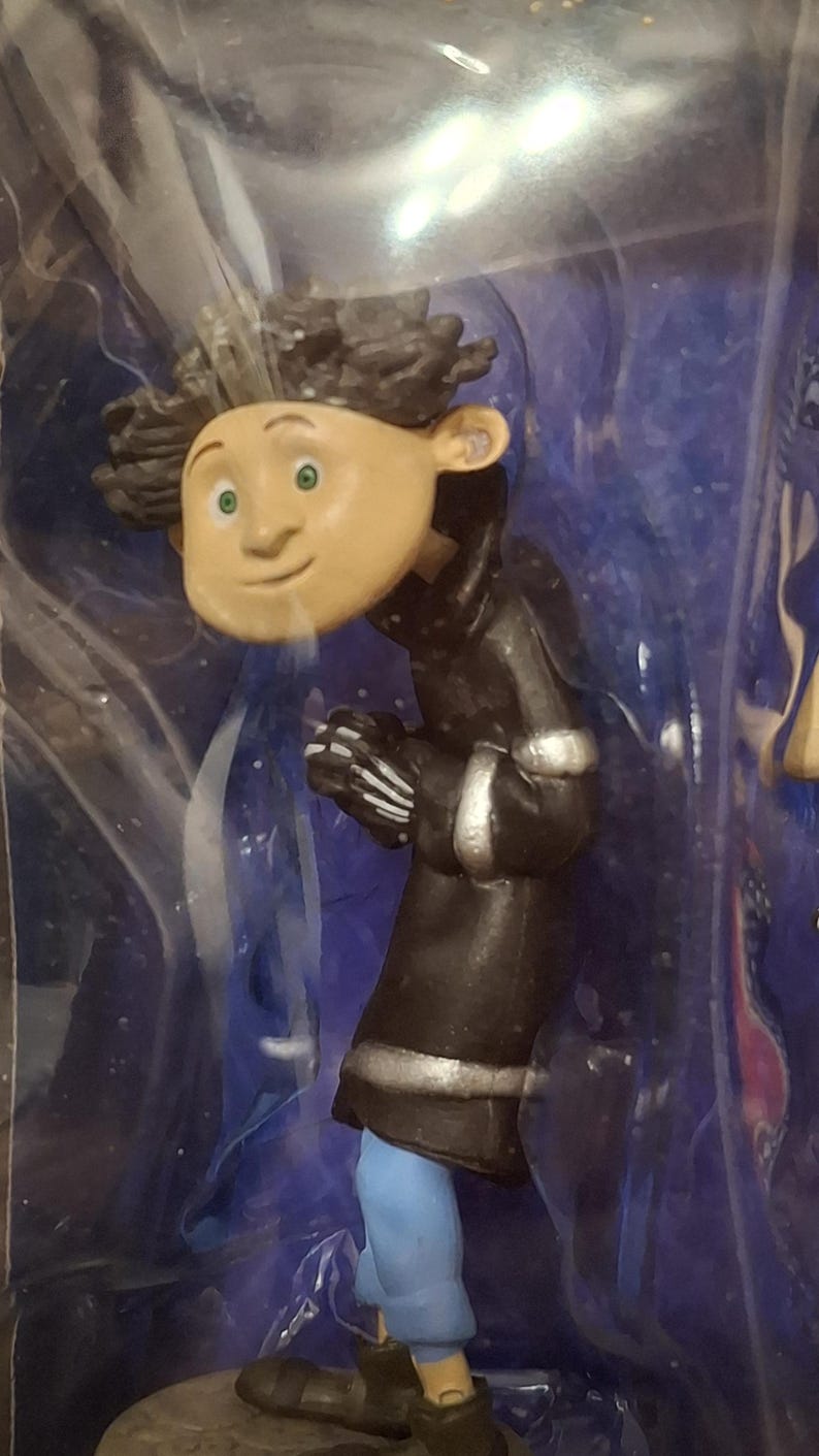 K&ouml;nnte beinhalten: Eine Figur einer Figur aus dem Animationsfilm "Coraline". Die Figur hat einen hellen Teint, gr&uuml;ne Augen und dunkles, lockiges Haar. Sie tr&auml;gt einen schwarzen Mantel mit silbernen Verzierungen, eine blaue Hose und schwarze Schuhe.