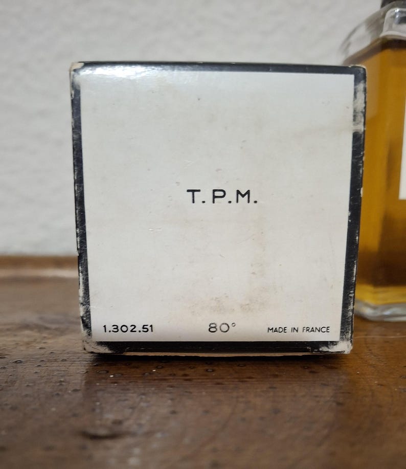 Pode incluir: Close-up de uma caixa de perfume vintage com o texto "T.P.M." em preto. Abaixo, os n&uacute;meros "1.302.51" e "80&deg;", com "MADE IN FRANCE" impresso na parte inferior. A caixa &eacute; branca com uma borda preta.