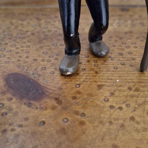 K&ouml;nnte beinhalten: Eine kleine Figur einer Person in einem schwarzen Anzugsakko und passender schwarzer Hose. Die Figur hat hellh&auml;utige H&auml;nde, tr&auml;gt braune Schuhe und h&auml;lt einen dunkelbraunen Gehstock in einer Hand. Die Farbe weist leichte Abnutzungen auf.