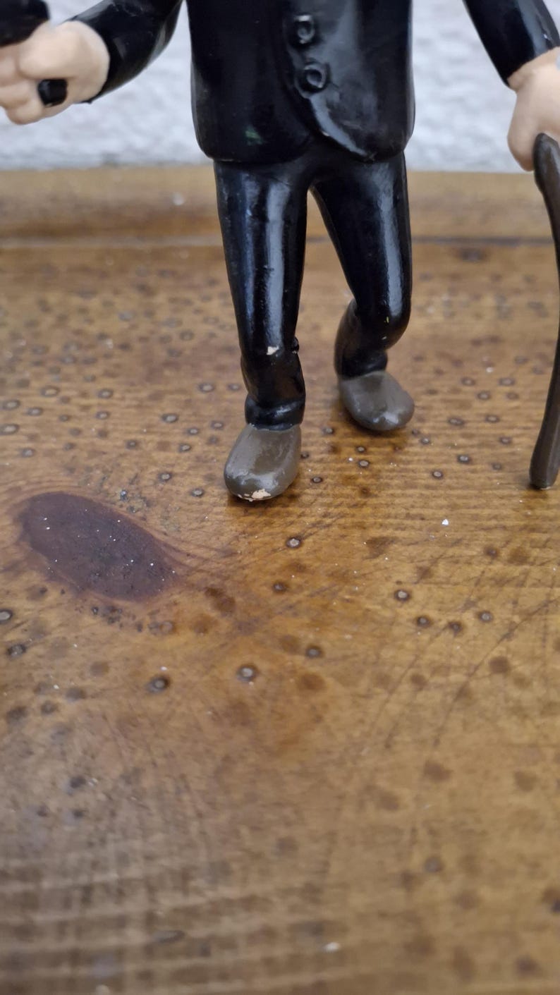 K&ouml;nnte beinhalten: Eine kleine Figur einer Person in einem schwarzen Anzugsakko und passender schwarzer Hose. Die Figur hat hellh&auml;utige H&auml;nde, tr&auml;gt braune Schuhe und h&auml;lt einen dunkelbraunen Gehstock in einer Hand. Die Farbe weist leichte Abnutzungen auf.