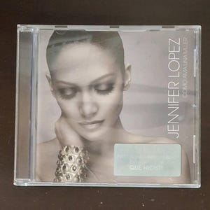 Op de afbeelding: Een muziek-cd in een doorzichtig plastic doosje met het album Como Ama Una Mujer van Jennifer Lopez. De hoes toont een zwart-witportret van de artiest met een armband met textuur, met de tekst Jennifer Lopez verticaal op de hoes gedrukt.