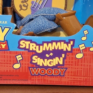 Puede incluir: Una caja de la mu&ntilde;eca "Strummin' Singin' Woody" de Toy Story 2. La caja es azul y roja con el logotipo de Disney Pixar. El texto "Strummin' Singin' Woody" est&aacute; en morado y blanco. Incluye pilas. Para mayores de 4 a&ntilde;os.