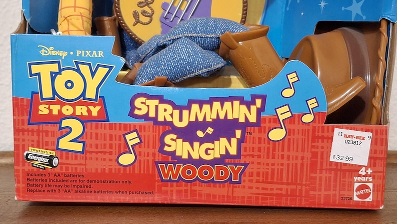 Puede incluir: Una caja de la mu&ntilde;eca "Strummin' Singin' Woody" de Toy Story 2. La caja es azul y roja con el logotipo de Disney Pixar. El texto "Strummin' Singin' Woody" est&aacute; en morado y blanco. Incluye pilas. Para mayores de 4 a&ntilde;os.