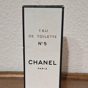 Pode incluir: Caixa de perfume retangular com frente branca e borda preta. A caixa apresenta o texto "EAU DE TOILETTE N&deg;5 CHANEL PARIS" em letras pretas. A caixa est&aacute; sobre um fundo neutro.