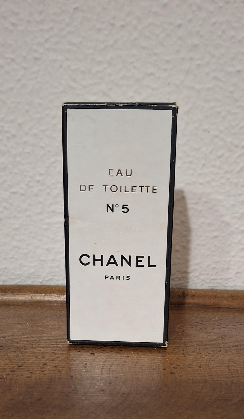 Pode incluir: Caixa de perfume retangular com frente branca e borda preta. A caixa apresenta o texto "EAU DE TOILETTE N&deg;5 CHANEL PARIS" em letras pretas. A caixa est&aacute; sobre um fundo neutro.