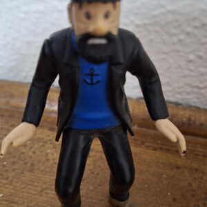 K&ouml;nnte beinhalten: Eine kleine Figur eines b&auml;rtigen Kapit&auml;ns, der eine schwarze M&uuml;tze mit gelbem Emblem, eine schwarze Jacke, einen blauen Pullover mit einem schwarzen Ankersymbol auf der Brust, eine schwarze Hose und hellbraune Schuhe tr&auml;gt und aufrecht steht.