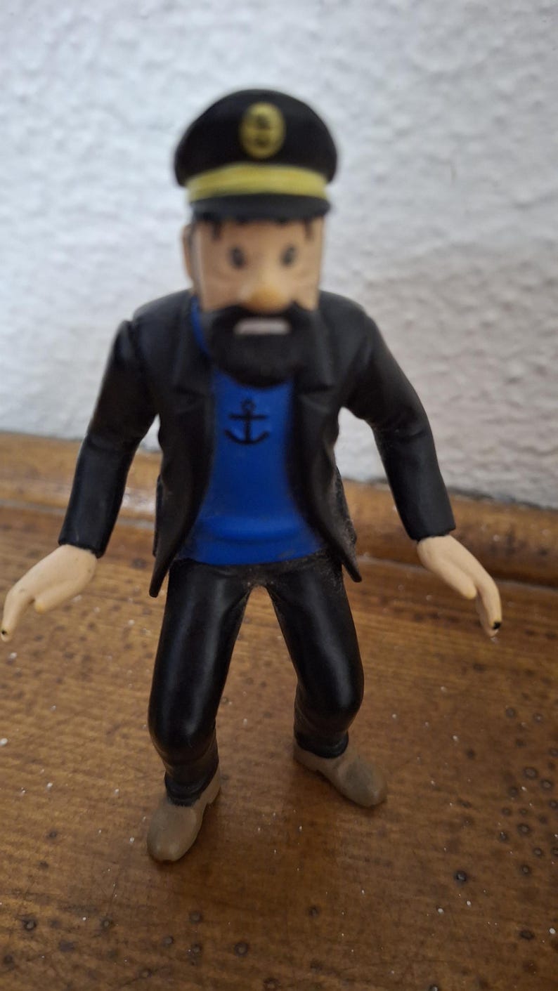 K&ouml;nnte beinhalten: Eine kleine Figur eines b&auml;rtigen Kapit&auml;ns, der eine schwarze M&uuml;tze mit gelbem Emblem, eine schwarze Jacke, einen blauen Pullover mit einem schwarzen Ankersymbol auf der Brust, eine schwarze Hose und hellbraune Schuhe tr&auml;gt und aufrecht steht.