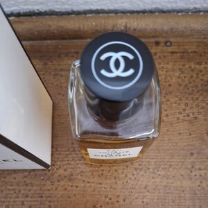 Pode incluir: Um frasco de perfume de vidro transparente com uma tampa preta com um logotipo branco. O frasco cont&eacute;m um l&iacute;quido &acirc;mbar claro. Uma caixa branca com texto preto &eacute; parcialmente vis&iacute;vel &agrave; esquerda.