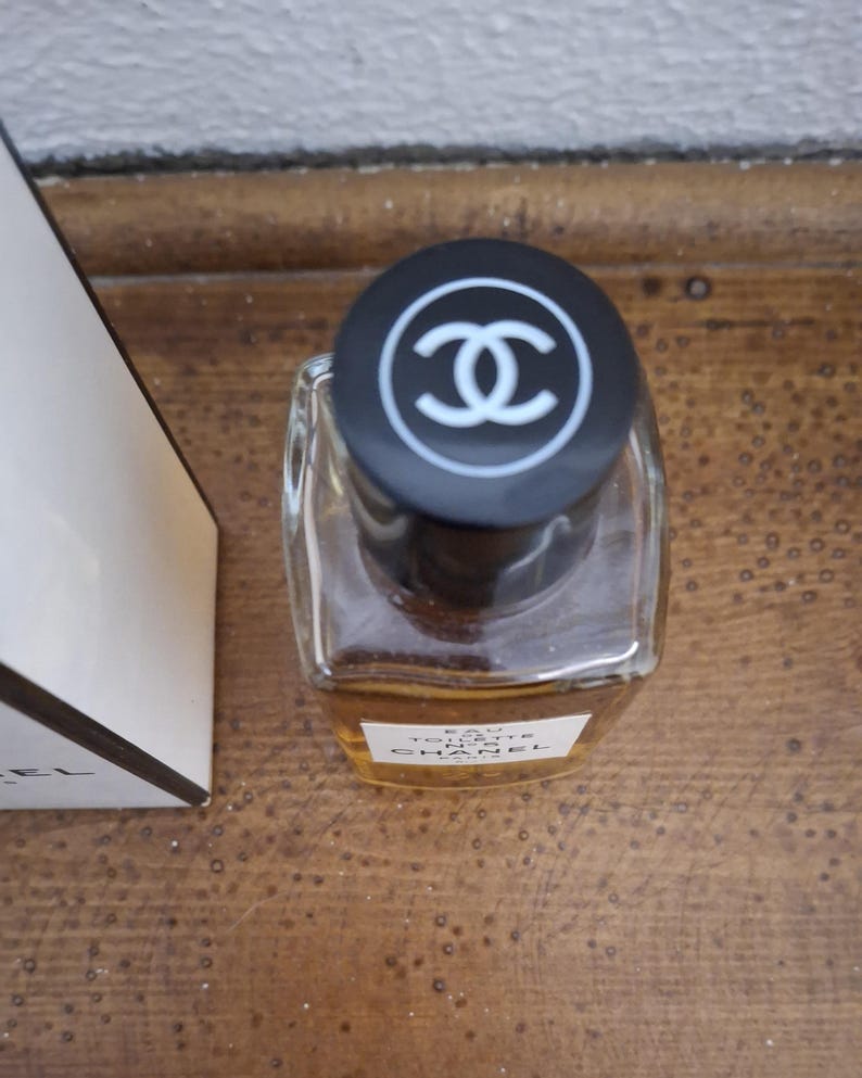 Pode incluir: Um frasco de perfume de vidro transparente com uma tampa preta com um logotipo branco. O frasco cont&eacute;m um l&iacute;quido &acirc;mbar claro. Uma caixa branca com texto preto &eacute; parcialmente vis&iacute;vel &agrave; esquerda.