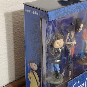 K&ouml;nnte beinhalten: Eine Box mit Figuren, die Charaktere aus dem Animationsfilm "Coraline" zeigen. Die Box ist blau mit Charakterillustrationen und dem Text "Ages 14 & Up". Die Figuren sind durch ein transparentes Kunststofffenster sichtbar.