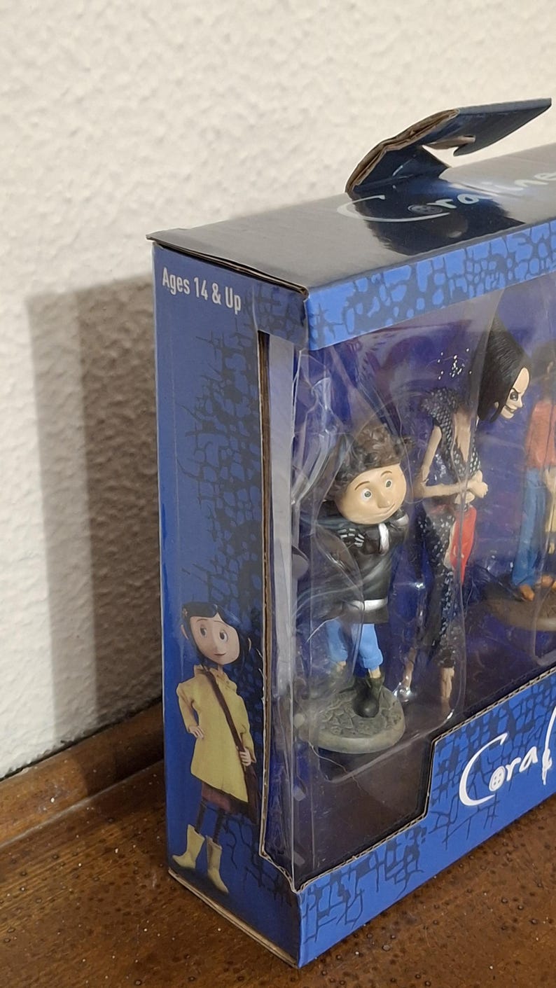 K&ouml;nnte beinhalten: Eine Box mit Figuren, die Charaktere aus dem Animationsfilm "Coraline" zeigen. Die Box ist blau mit Charakterillustrationen und dem Text "Ages 14 & Up". Die Figuren sind durch ein transparentes Kunststofffenster sichtbar.