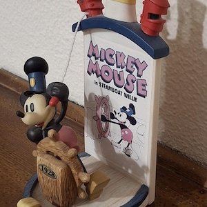 May include: A Steamboat Willie Mickey Mouse figurine. The figurine features Mickey Mouse at the helm of a ship, with the text "Mickey Mouse in Steamboat Willie." The figurine has a blue, white, and red color scheme.