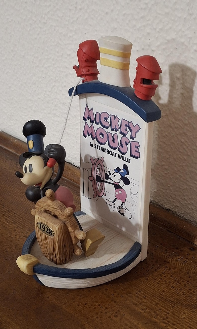 May include: A Steamboat Willie Mickey Mouse figurine. The figurine features Mickey Mouse at the helm of a ship, with the text "Mickey Mouse in Steamboat Willie." The figurine has a blue, white, and red color scheme.