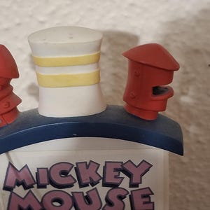 May include: A Mickey Mouse themed decorative item. The item features a white smokestack with yellow stripes, red accents, and the words "Mickey Mouse" in purple and white. The item has a blue base.